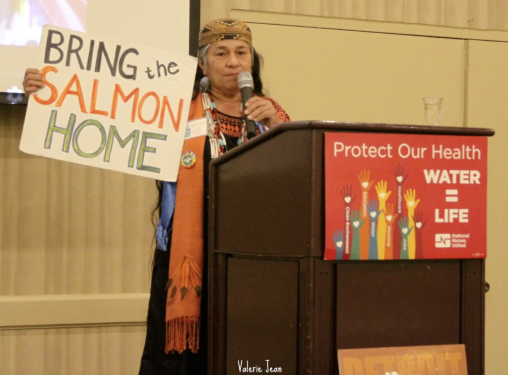 A speaker holds a sign that reads, "Bring the salmon home" in front of a podium with a sign that reads, "Water = Life."